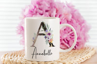 A white mug sat on a wicker coaster with bright pink blurry flowers in the background. On the mug is a beautiful large capital A with purple and lavender flowers. Below the large letter is the name Annabelle written is a stylish calligraphy font.
