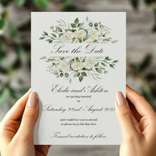 A person holds a vertical white wedding save-the-date card with watercolor floral designs in green and white, with elegant black script text. The background is a softly blurred outdoor setting with green foliage and gray stone.