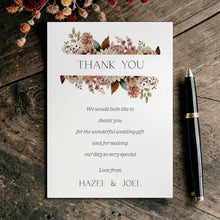 A wedding thank-you card with floral watercolor designs at the top and elegant black text on a white background, styled on a rustic dark wood surface next to a black pen.