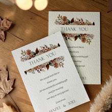 Two wedding thank-you cards with floral watercolor designs are arranged on a wooden surface with scattered autumn leaves and warm bokeh lights in the background.