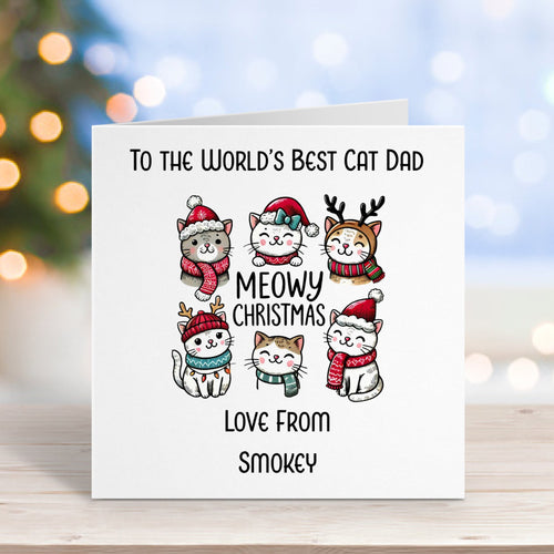Card with cartoon cats wearing Christmas hats on a wooden surface with blurred lights in the background