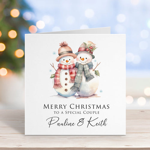 Christmas card with two snowpeople wearing scarves and hats on a blurred Christmas tree background.