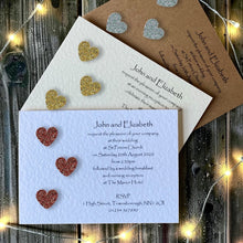 A coordinated wedding invitation suite arranged on a rustic wooden surface with warm string lights. The set includes a light blue invitation card with glittery red, gold, and copper hearts, a white invitation card with gold glitter hearts, and a kraft brown envelope with silver glitter hearts.