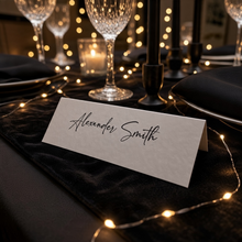 A white paper place card with the name 'Alexander Smith' written in black script, set on a black table runner surrounded by warm string lights and a dinner table setting.