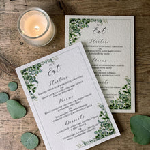 Two printed wedding stationery cards with green foliage borders and elegant script text, arranged on a rustic wooden surface beside a lit candle and green leaves. The front card is slightly overlapping the second card behind it.