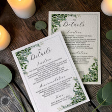 Two Esme wedding information cards with sage eucalyptus corners, fine borders and black script headings, displayed on wood beside candles and greenery