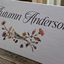 A close-up of a white personalized wooden sign with the name 'Autumn Anderson' in black cursive script and a watercolor-style autumn floral branch across the lower middle, resting on a rustic wooden surface.