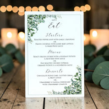 A vertical wedding menu card with elegant black script text on a white background, decorated with green eucalyptus leaves at the top and bottom corners, displayed on a rustic wooden table with warm bokeh lights in the background.