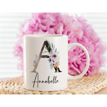"Annabelle" Floral Initial personalised mug gift. A white mug sat on a wicker coaster with bright pink blurry flowers in the background. On the mug is a beautiful large capital A with purple and lavender flowers. Below the large letter is the name Annabelle written is a stylish calligraphy font.